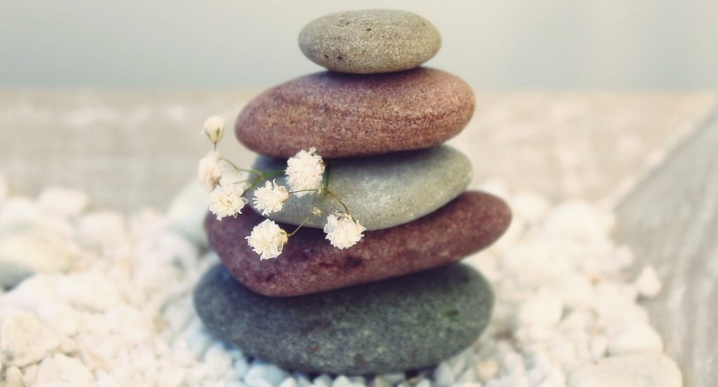 Five different colored rocks are stacked on top of each other, balancing on a bed of small white stones. A sprig of Baby's Breath is peeking out from the middle of the stack. The stones get smaller toward the top. 